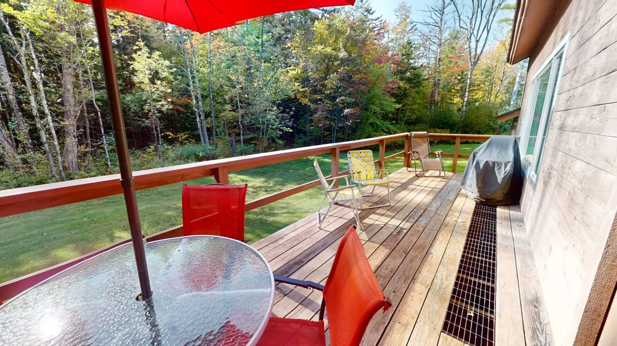 A cheerful, relaxed exterior view highlighting a wooden deck with a red umbrella, a glass table for outdoor dining, a green lawn, and surrounding trees. The scene evokes the pleasures of al fresco meals and time spent outdoors.
