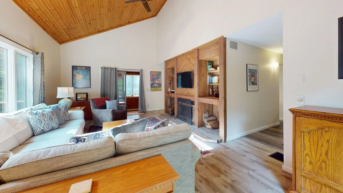 The image shows a warm, inviting living room with a plush sofa adorned with patterned cushions, a rustic wooden coffee table, a glowing fireplace, expansive windows offering scenic views, and a bookshelf filled with books. Sunlight streams in, creating a cozy, welcoming atmosphere perfect for relaxation and gathering.