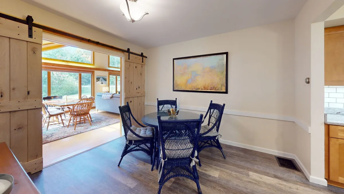 A bright dining area with a round dining table, blue wicker chairs, a sliding barn door, and large windows that let in plenty of natural light. The arrangement invites shared meals, stories, and relaxed gatherings by the window.