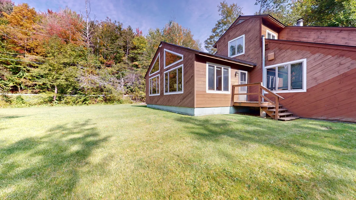 The image features the wooden exterior of the property, highlighted by large windows and a landscaped yard. The inviting yard offers a perfect play area for children and a relaxing spot for parents to enjoy the surrounding Vermont nature. The setting suggests opportunities for family-friendly outdoor gatherings and activities.