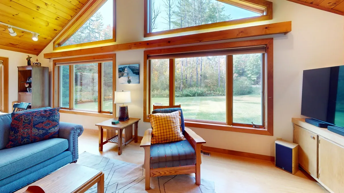 A bright, inviting living room with large windows allowing natural light to flood the space, featuring a comfortable seating area with a plush sofa, decorative pillows, a TV, and warm wooden accents. The view outside brings an extra layer of tranquility and connection to nature.