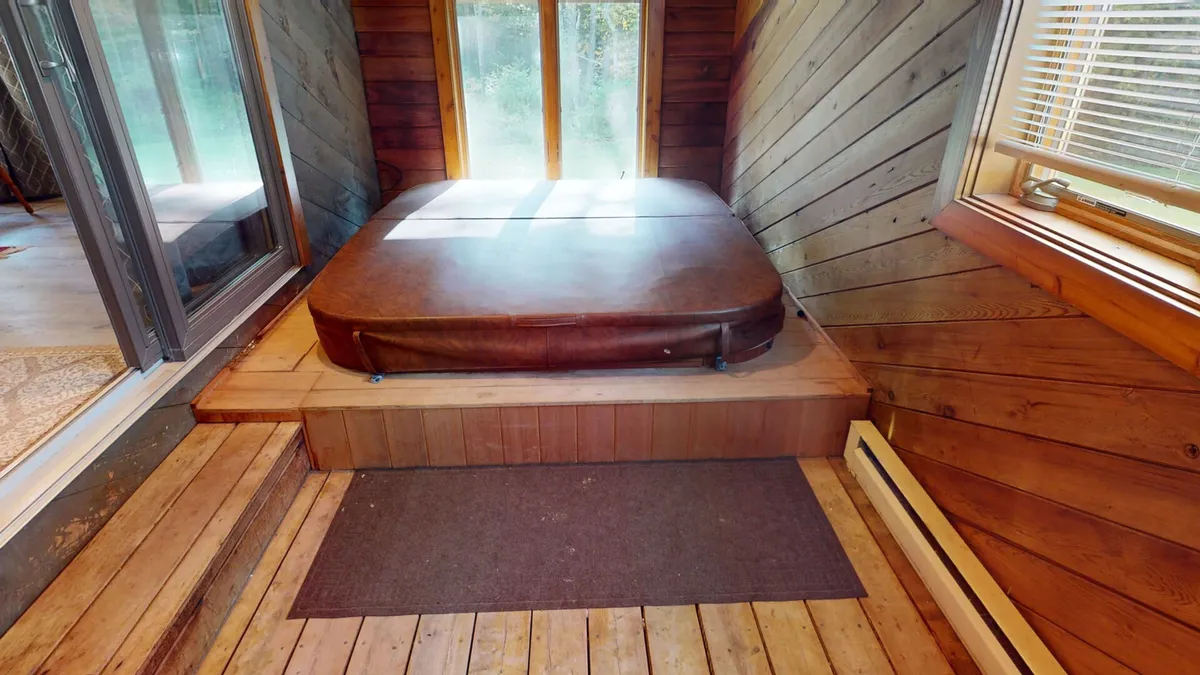 This interior image centers on a luxurious hot tub set against wooden walls, with large windows that invite natural light and display tranquil views of surrounding nature. The steam hinting at warmth, coupled with the serene atmosphere, highlights the ultimate private spa experience. Subtle decor enhances the relaxing, upscale vibe, and the room feels removed from the rest of the home for an exclusive, retreat-like ambiance.