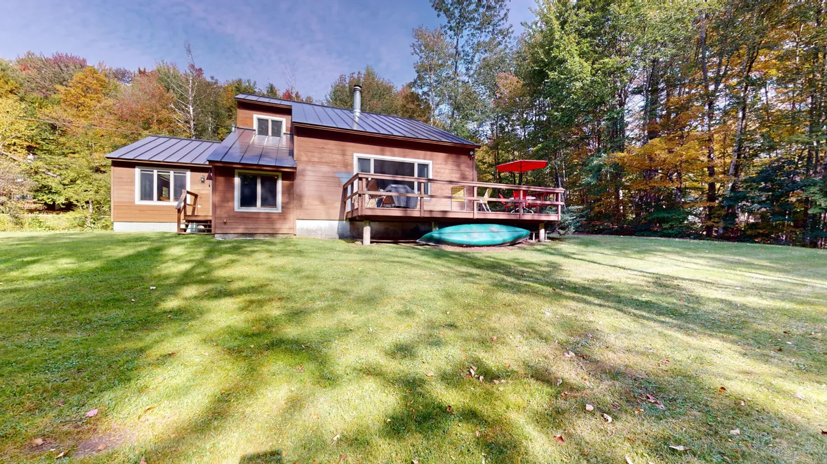 This inviting exterior scene showcases a wooden house with a spacious deck featuring a red umbrella, green kayak, and an expansive green lawn. The space is ideal for outdoor gatherings, barbecues, and relaxing after a day of adventure.