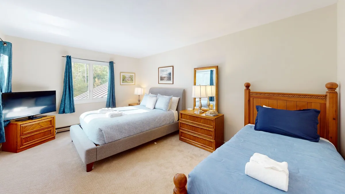 A warmly lit bedroom featuring a queen-sized bed with light blue bedding and a twin bed with a dark blue pillow, complemented by a wooden nightstand with lamps, a television on a dresser, and framed pictures on the wall. The window allows natural light to stream in, creating a cozy and inviting atmosphere suitable for relaxing or enjoying breakfast in bed.
