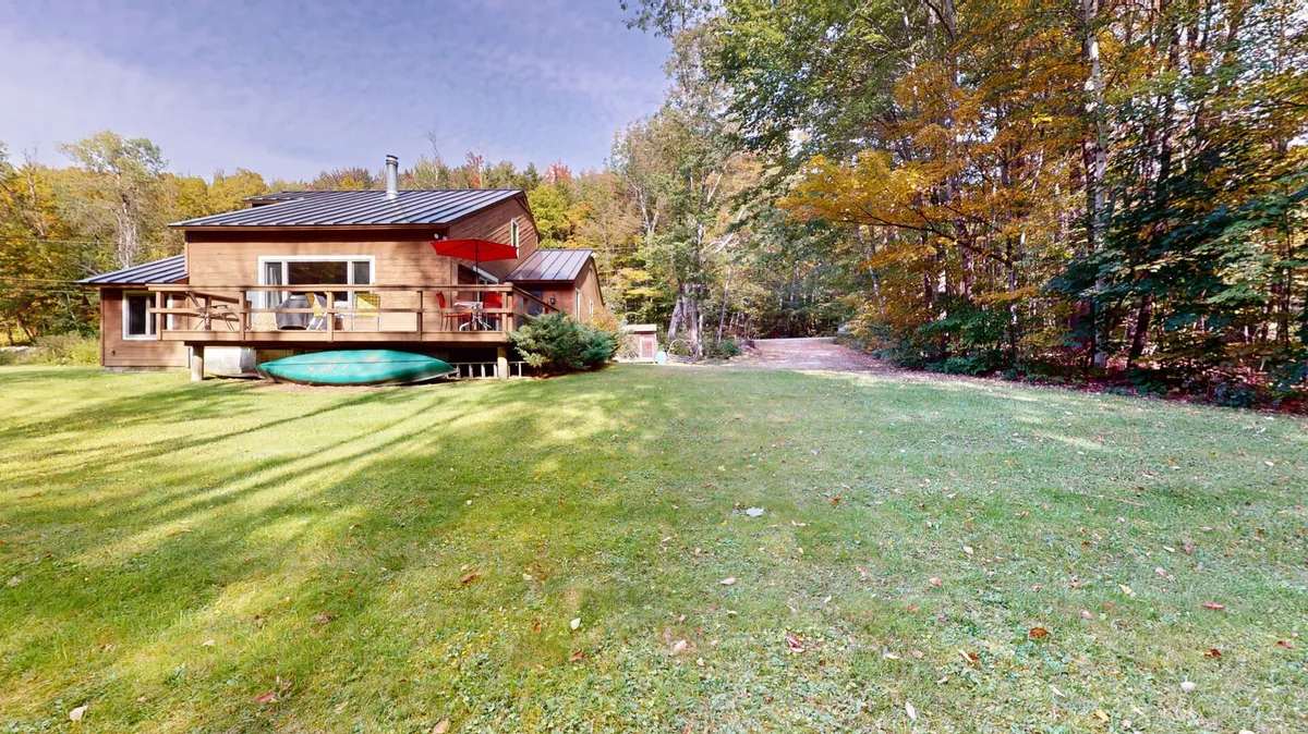 A serene exterior shot of a wooden house surrounded by a lush green lawn and trees. A green kayak rests on the lawn, suggesting opportunities for outdoor adventure. The peaceful setting reflects the natural beauty and recreational spirit of the area.