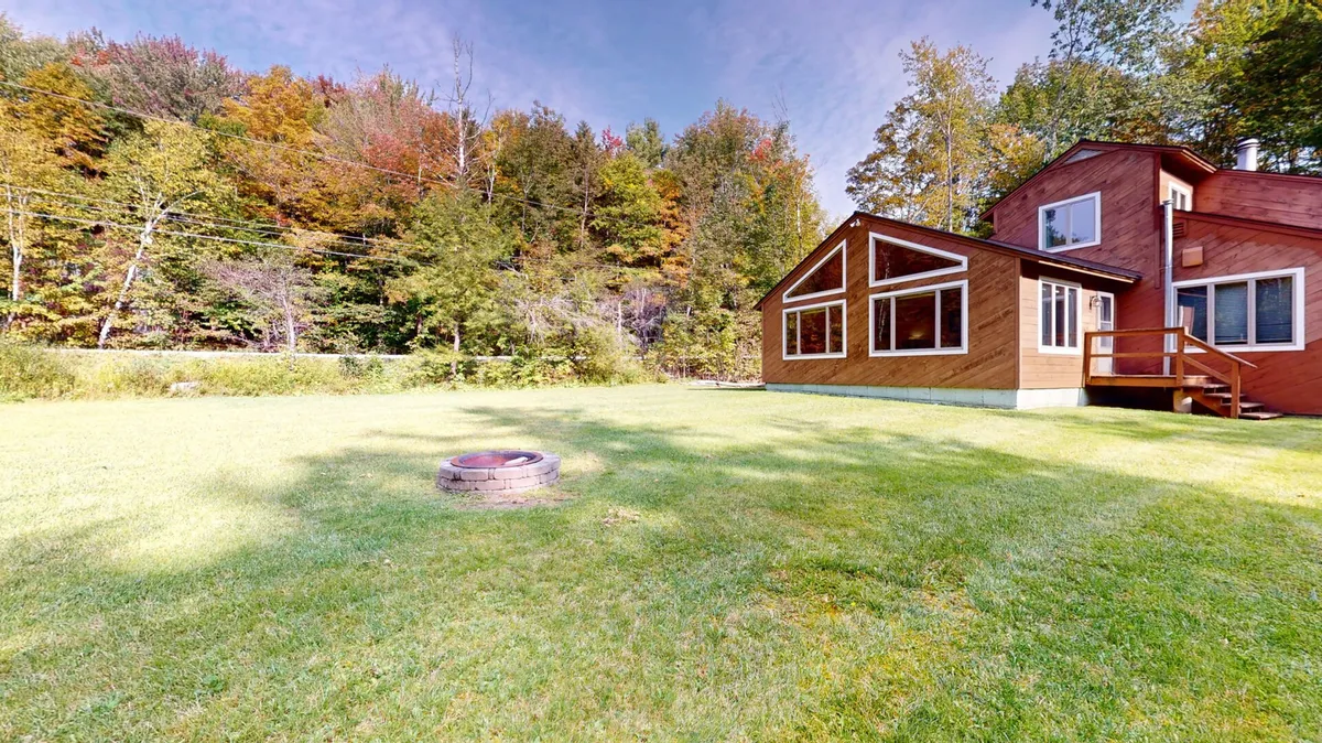 This image showcases the exterior of the property: a charming house surrounded by a lush lawn and tree line, featuring a cozy firepit area. The scene evokes a sense of relaxation and connection to nature, with the home nestled in a peaceful, wooded setting perfect for unwinding after a day of winter sports.
