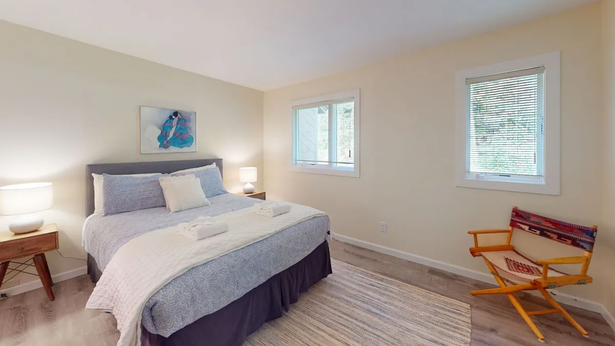 A serene bedroom highlighted by a comfortable bed dressed in soft linens, tasteful artwork above the headboard, windows that let in gentle daylight, and modern lighting fixtures. A cozy chair in the corner invites quiet moments of reading or relaxation, reinforcing the tranquil vibe of a master suite retreat.